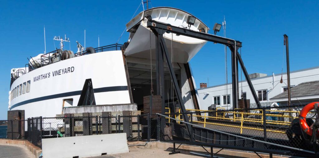A close-up view of the Martha's Vineyard ferry, with the ramp down at the dock, ready to load passengers and vehicles. The bright blue sky and surrounding structures emphasize the island's welcoming vibe and transport accessibility.